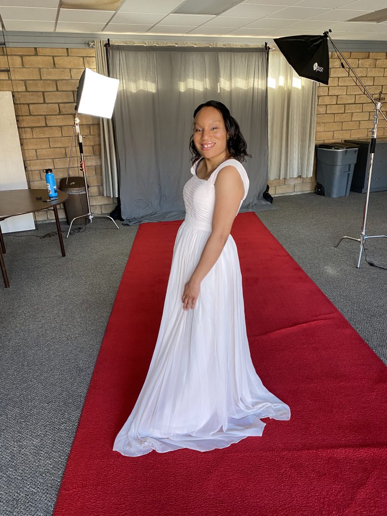 Saida Mahoney, a rare disease advocate, wearing a white dress and smiling for a photo on a red carpet with a gray curtain in the background for the California Miss Amazing pageant.